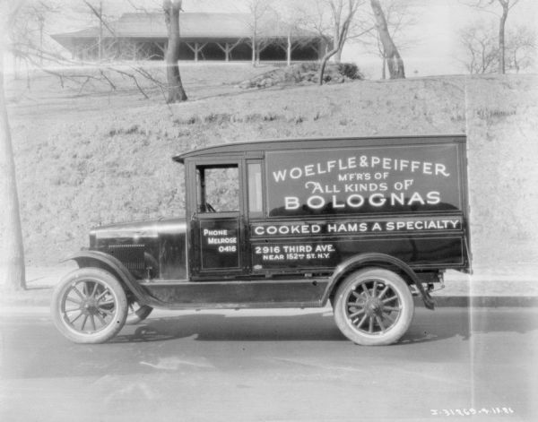 Left side view of a delivery truck parked along a curb. A sign on the truck reads: "Woelfle & Peiffer M'F'R'S of All Kinds of Bolognas." There is a pavilion at the top of a steep hill in the background.
