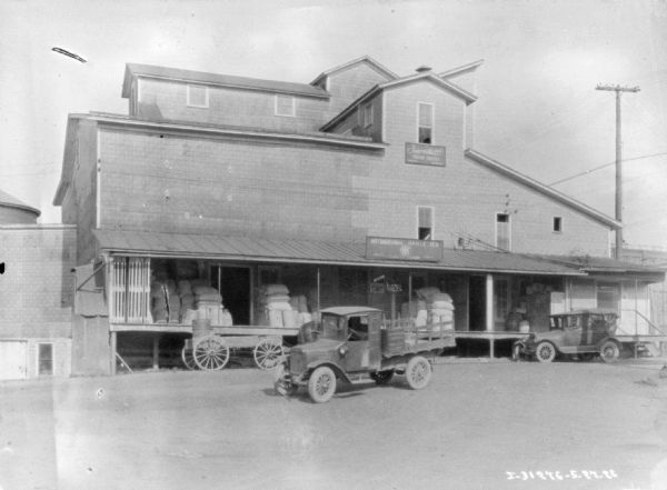 Exterior view of a truck parked near a dealership. There is a wagon parked near a porch or loading dock, which is stocked with sacks piled under the roof.