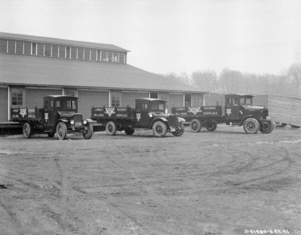 View across parking area towards lumber delivery trucks parked in front of a large, wooden sided building, with clerestory windows. A sign on the side of the trucks reads: "Proctor Mfg. Co."