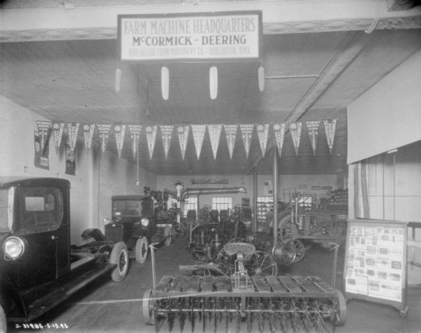 Interior view of banners, pennants, literature, trucks, and agricultural implements on display at a dealership. A large sign attached to a beam on the ceiling reads: "Farm Machine Headquarters, McCormick-Deering, Burlington Farm Machinery Co., Burlington, Iowa." Pennants hanging in a line just below the ceiling read: "McCormick Deering, Farm Equipment," and "McCormick Deering Ball Bearing Separators."