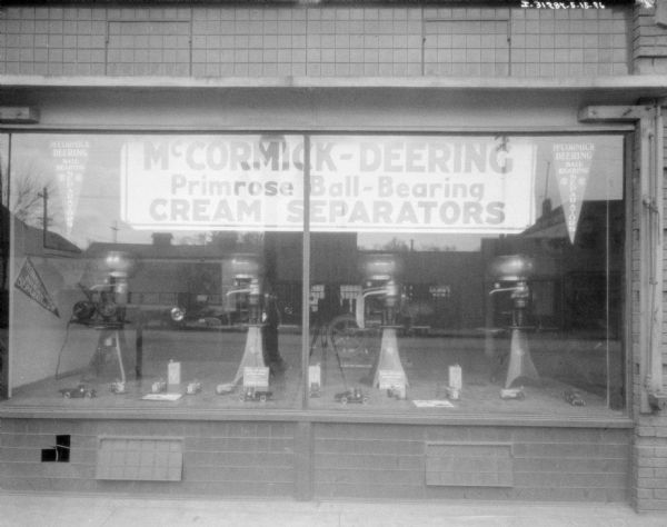 Outdoor view of a show window with a banner inside reading: "McCormick-Deering Primrose Ball-Bearing Cream Separators." On display are four cream separators, along with toy trucks and tractors on the floor in front of them.