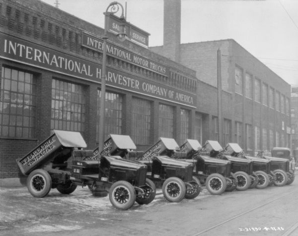 Six dump trucks are parked at an angle in front of a factory building. The dump truck beds are tipped up. Signs on the factory reads: "International Harvester Company of America," and "International Motor Trucks." Painted on the side of the dump trucks is a sign that reads: "W.J. Gallagher, Contracting."