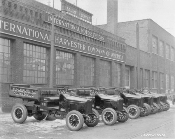Six dump trucks are parked at an angle in front of a factory building. Signs on the factory read: "International Harvester Company of America," and "International Motor Trucks." Painted on the side of the dump trucks is a sign that reads: "W.J. Gallagher, Contracting."