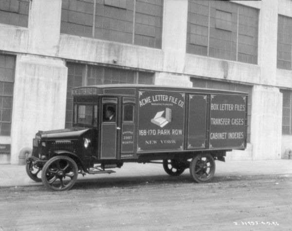 A man is sitting in the driver's seat of a delivery truck is parked along the side of an industrial building. Painted on the side and front of the truck are signs that read: "Acme Letter File Co., Manufacturers, 168-170 Park Row, New York."