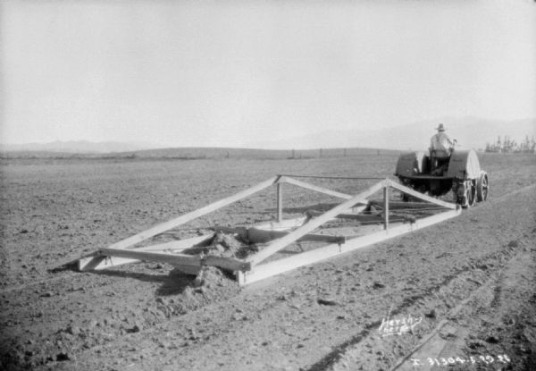 Three-quarter view from right rear of a man driving a tractor to pull a frame/ground leveler in a field.