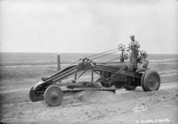 Left side view of a man driving a grader. Painted on the side: "Austin Motor Grader."