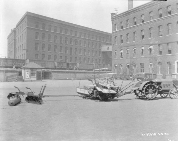 View across factory yard towards a tractor hooked up to a harvester and another agricultural implement. There are railroad cars on elevated tracks in the background near another factory building.
