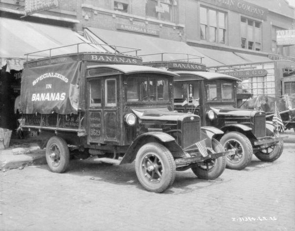 Delivery trucks, with American flags attached to the hood, are backed up to storefronts. Signs on the truck read: "Specializing in Bananas." A sign above the awning of one of the storefronts reads: "A. Huizinga & Sons."