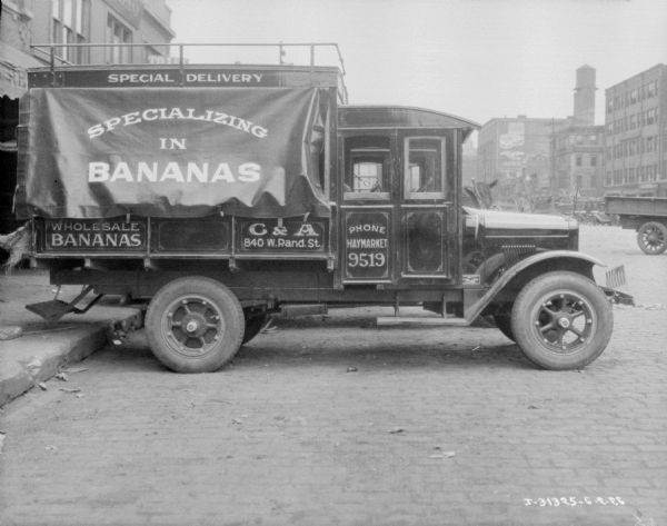 Delivery trucks, with American flags attached to the hood, are backed up to storefronts. Signs on the truck read: "Specializing in Bananas."