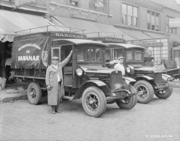 Two men are posing with delivery trucks backed up to storefronts. Signs on the truck read: "Specializing in Bananas." A sign above the awning of one of the storefronts reads: "A. Huizinga & Sons."