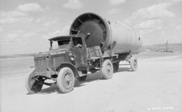 View of a driver sitting in the driver's seat of a truck on a highway in the western U.S. The open truck bed is loaded with large parts. Oil wells are in the background.