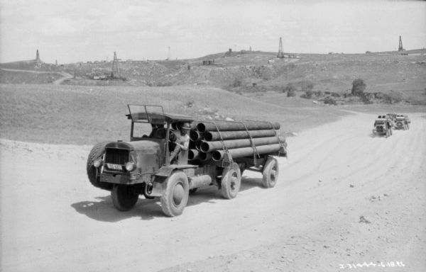 View of a driver sitting in the driver's seat of a truck on a highway in the western U.S. The open truck bed is loaded with large parts. Oil wells are in the background.