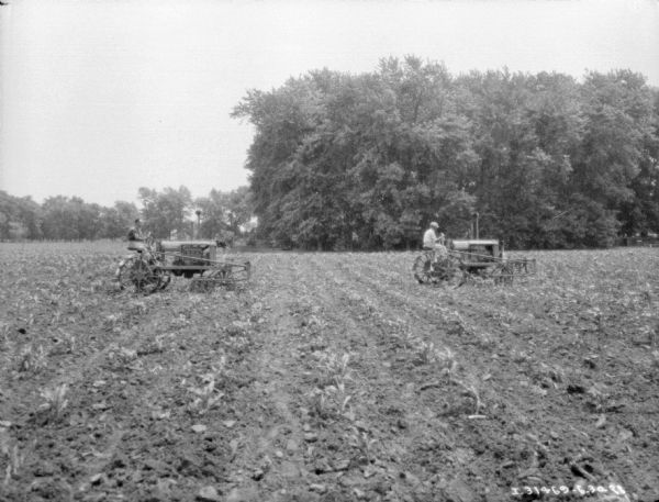 View across field towards two men with Farmall tractors cultivating a field.