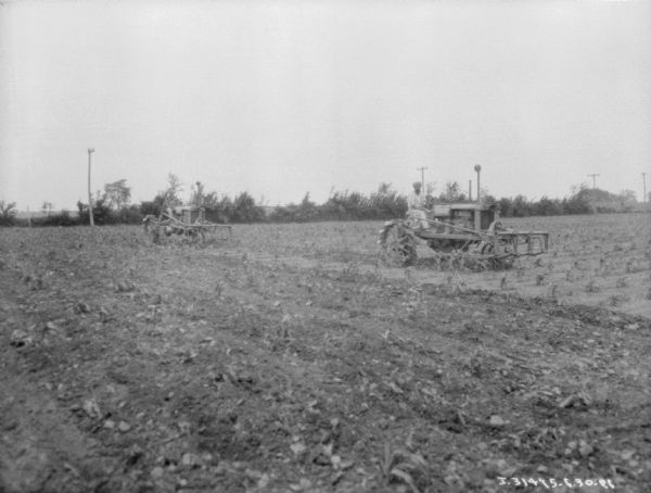 View across field towards two men with Farmall tractors cultivating a cornfield.