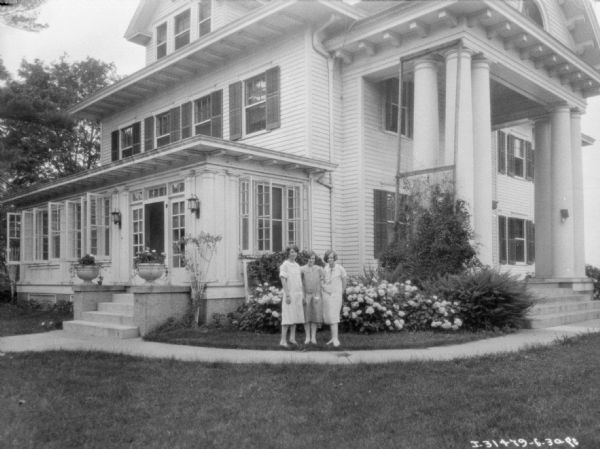 View across lawn towards three women posing in front of a large home with a manicured lawn.