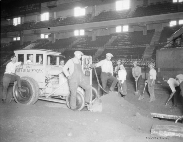 Group of men working on the grounds of a baseball stadium. One man is sitting in a truck which has a painted sign on the side that reads: "D.S.C. City of New York." 
