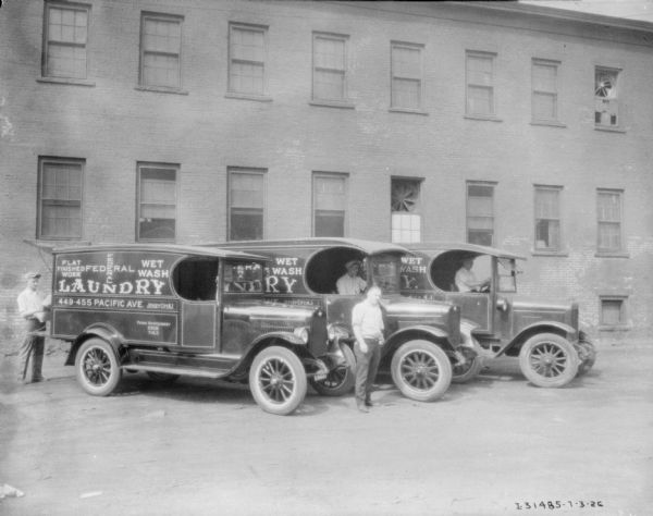View of four men posing with three delivery trucks for Federal Laundry. There is a large brick building in the background.