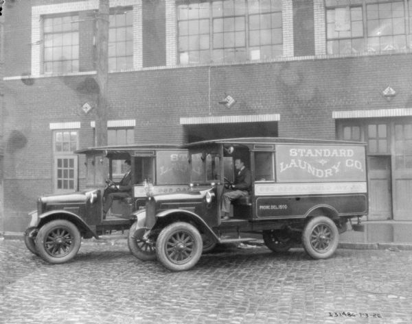 View across street towards two men sitting in the driver's seats of two laundry delivery trucks. The trucks are parked at an angle in front of a brick building. The signs on the truck read: "Standard Laundry Co."