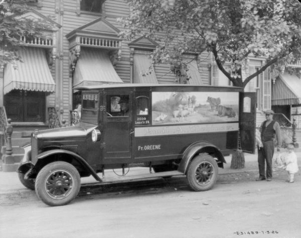 A man is sitting in the driver's seat of a delivery truck parked along a curb. A man is standing at the back of the truck holding a chicken. There is a young girl standing next to him. In the background a woman is sitting on the stoop of a wood building with awnings over the windows. The truck has a sign that includes an illustration of a person leading sheep and chickens down a path in a rural area. The sign on the truck reads: "Emil A. Fichtner."