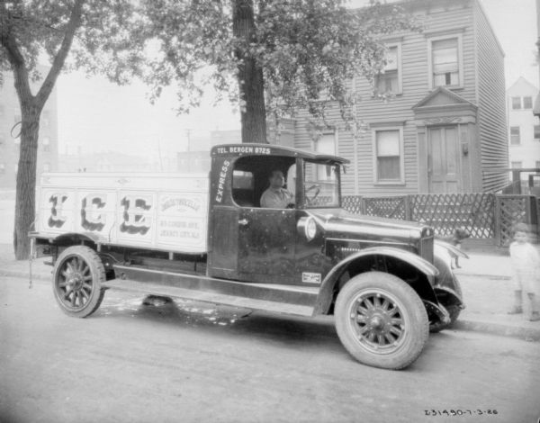 Passenger side view of a man sitting in the driver's seat of a truck parked on a street. There is a dog just behind the truck, and a young child standing on the terrace. There is a house in the background. The sign on the truck reads: "Ice, Express."