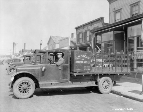 View down street towards a man sitting in the driver's seat of a truck backed up to a sidewalk in front of a storefront. The sign on the truck reads: "J.H. Stout, Hampton Bays LI."