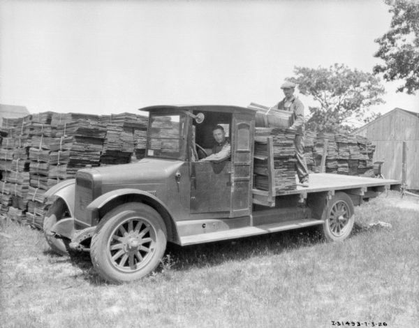 Two men in a lumber yard with a truck. One of the men is sitting in the driver's seat. The other man is standing in the truck bed, surrounded by stacks of lumber, and holding more lumber in his arms. Behind them are more stacks of lumber.