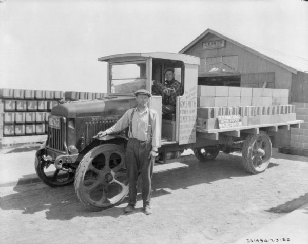 Two men with a C.W. delivery truck parked in front of a building for C.W. Smith. One man is standing and resting his arm on the fender, and the other man is sitting in the driver's seat. There is a load of cement blocks on the open bed of the truck. A sign painted on the side of the truck reads: "C.W. Smith, Power Tamp, Cement Blocks." A sign propped on the cement blocks reads: "International Motor Trucks, H.M. Reeve & Sons."