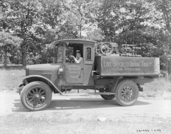 View of a young boy sitting in the driver's seat of a truck. There is someone sitting in the passenger seat behind him. The signs painted on the side of the truck read: "Live Speckled Brook Trout for Stocking," and "Pepperridge Lake Trout Hatchery, Eastport, L.I." There is equipment in the back of the truck bed.