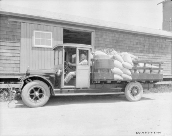 View of a man in the driver's seat of delivery truck. There are piles of sacks in the truck bed. In the background is a building with a large open door and a loading dock.