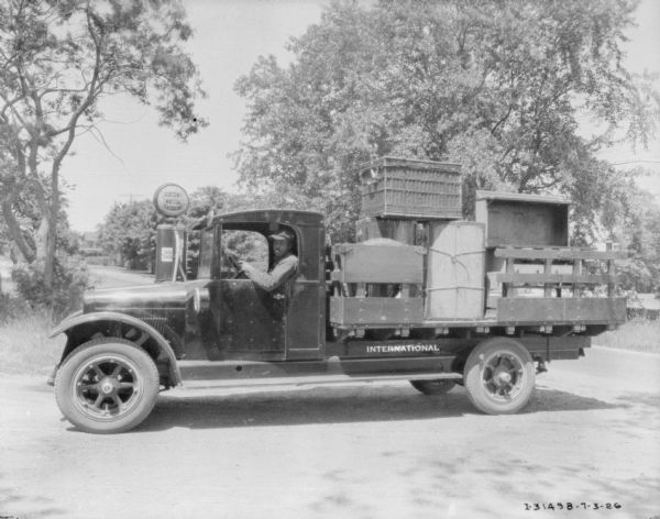 View of a man sitting in the driver's seat of a delivery truck. The back of the truck is loaded with items. There is a gas pump behind the truck, with a sign that reads: "Socony Motor Gasoline."