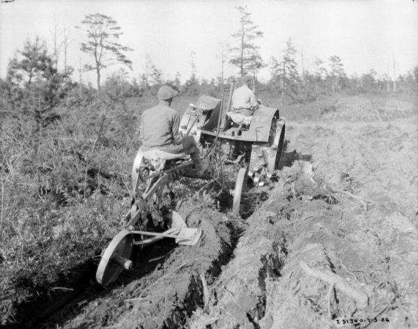 Rear view of a man riding a plow being pulled by a man on a 15-30 tractor in an overgrown area.