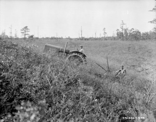 View along side of hill towards a man riding a plow being pulled by a man on a 15-30 tractor in an overgrown area.