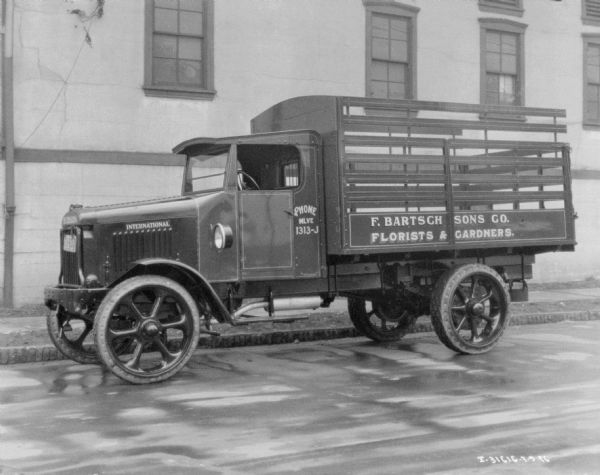 View across street towards a floral delivery truck parked along a curb in front of a building. The sign painted on the side of the truck bed reads: "F. Bartsch Sons Co. Florists & Gardners."