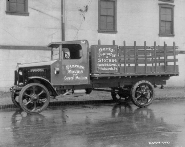 View across street towards a delivery truck parked along a curb in front of a building. The signs painted on the side of the truck bed read: "Derby Transfer & Storage," and "Storage, Moving General Hauling."
