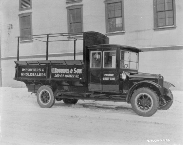 View across street towards a delivery truck parked along a curb in front of a building. There is snow on the ground. The sign painted on the side of the truck bed reads: "Importers & Wholesalers, I. Robbins & Son."