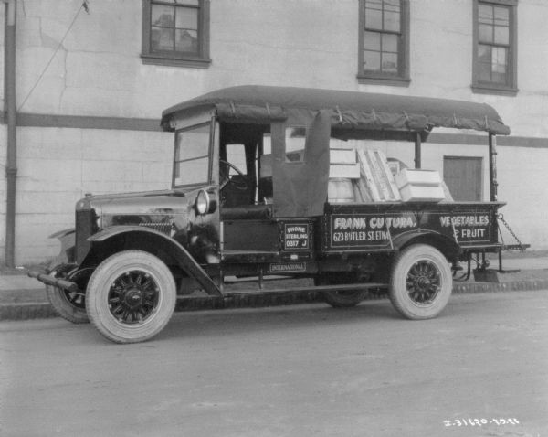 View across street towards a delivery truck parked along a curb in front of a building. The sign painted on the side of the truck bed reads: "Frank Cutura Vegetables & Fruit." There are crates piled in the truck bed. The canvas sides of the truck are rolled up above the side of the steering wheel, and the side of the truck bed. The section at the driver's side door is rolled down, and includes a window.