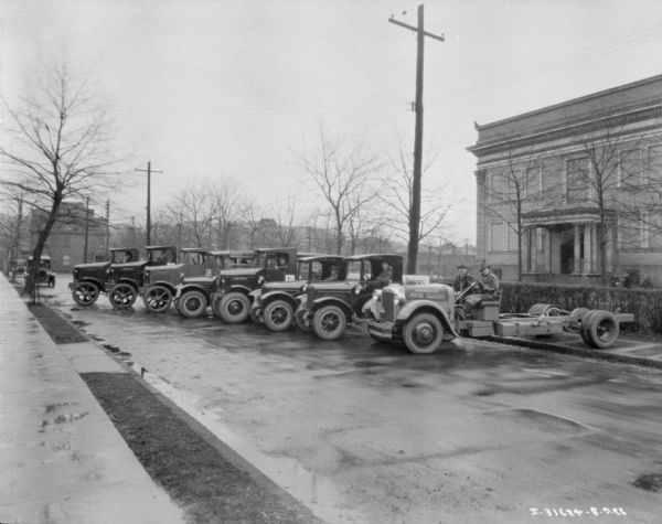 View from sidewalk towards a fleet of eight trucks parked at an angle in a street. There are men sitting in the driver's seat of each truck. There are two men sitting in an open truck on the far right which has no cab and no truck bed.