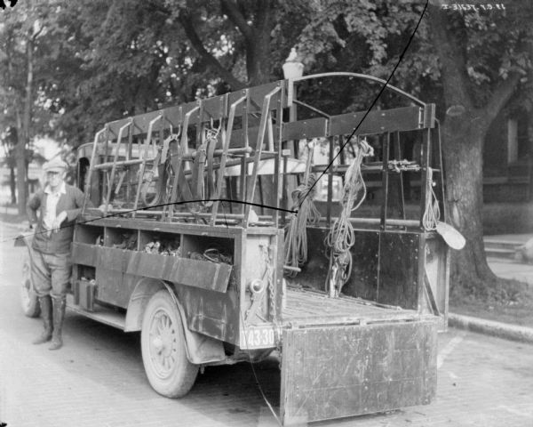 Three-quarter view from left rear of a man standing near the driver's side door of a truck. The back of the truck has open shelving on the side, and an open framework over the truck bed. Tools, ropes and other items are hanging from hooks on the side.