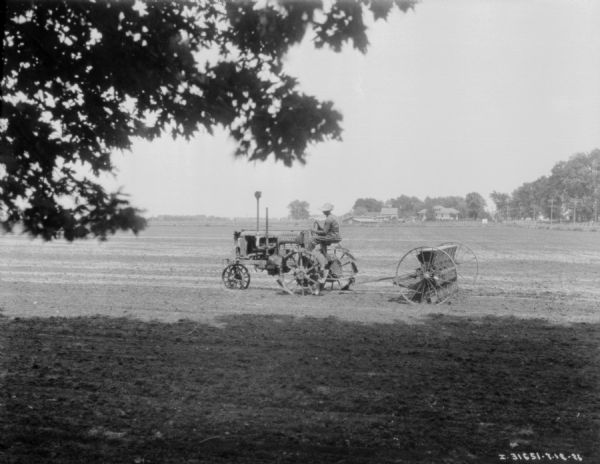 View of a man using a Farmall tractor to seed a field. There are farm buildings in the background.