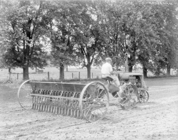 Three-quarter view from right rear of a man using a Farmall tractor to seed a field.