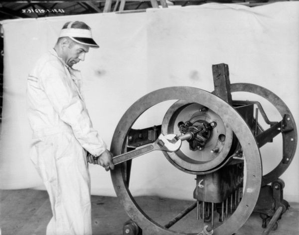 A man wearing a work coverall and a hat with a visor is working on machine parts. In the background is a white backdrop.