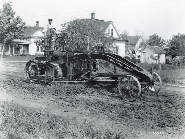 View of a man using an Austin Motor Grader on a road. There are houses in the background.