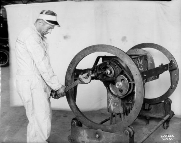 A man wearing a work coverall and a hat with a visor is tightening gears on a piece of large machinery. Behind him is a white backdrop.