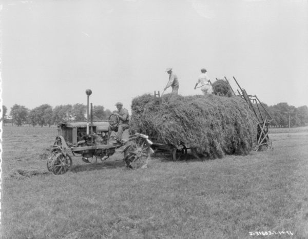 Three-quarter view from front left of a man driving a Farmall tractor pulling a wagon. Two men are standing on top of the hay and using pitchforks to pull hay from the hay stacker onto the wagon.