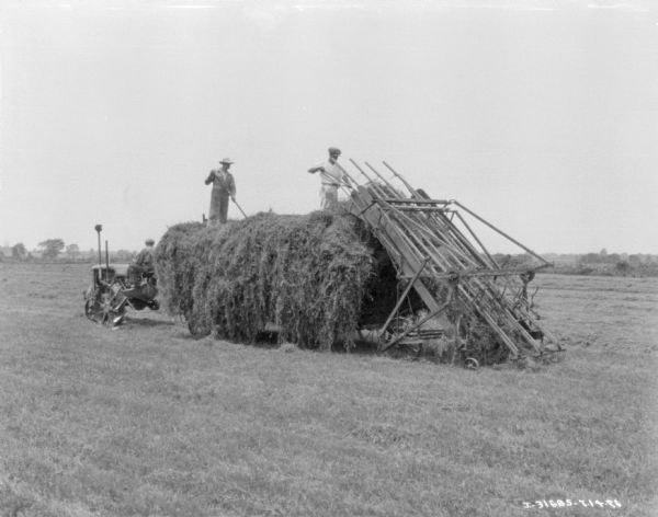 Three-quarter view from left rear of a man driving a Farmall tractor pulling a wagon. Two men are standing on top of the hay and using pitchforks to pull hay from the hay stacker onto the wagon.
