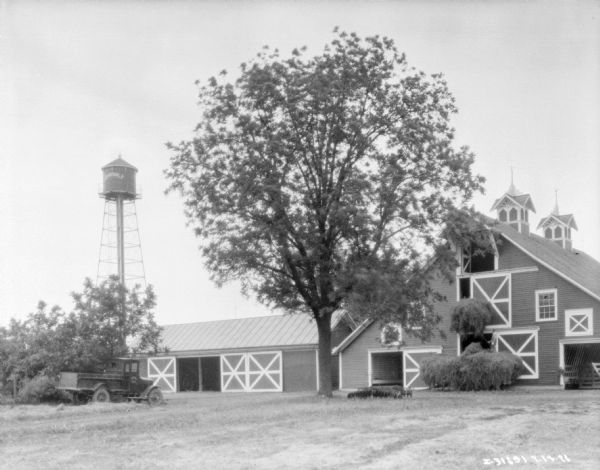 Farmer loading hay from a wagon into a large barn using a winch. There is another barn, a water tower, and a truck on the left.