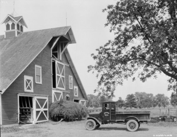 View of farmyard, where a man is standing on top of a large pile of hay on a wagon parked in front of a large barn. There is a truck parked nearby, and the sign on the driver's side door of the truck reads: "Forestdale Farm." 