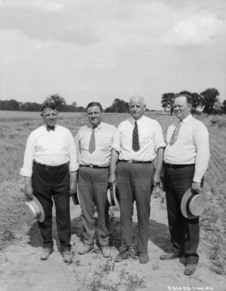 Four men are posing in a field, wearing white shirts, neckties and bow ties, and holding straw hats in their hands.