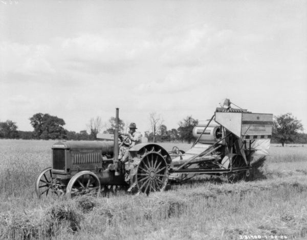 Three-quarter view from front left of a man driving a kerosene tractor pulling a McCormick-Deering No. 8 Power Drive Harvester Thresher.