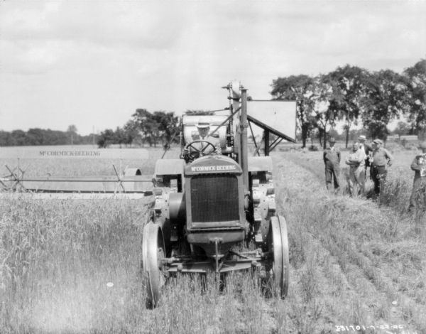 View from front of a man wearing a suit and hat driving a McCormick-Deering kerosene tractor pulling threshing equipment. A group of men are watching in the background on the right.